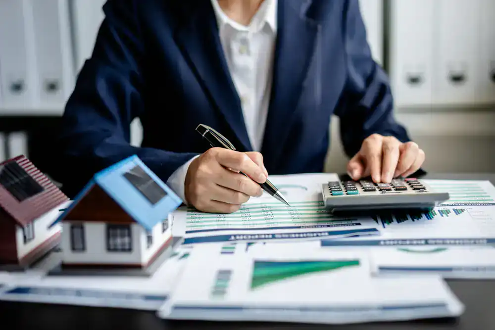 Realtor analyzing property documents with calculator and house models on desk.